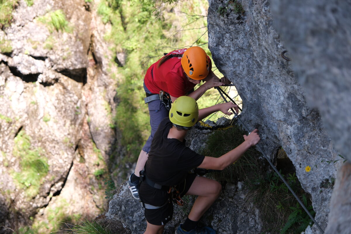 Zwei junge Männer klettern einen Klettersteig hinauf und halten sich an einem Stahlseil fest, mit Sicherungsleine, Helm und Gurten.