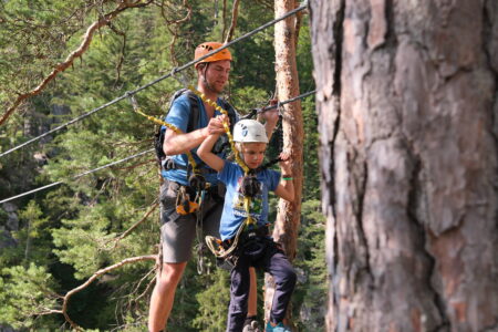 Holzhängebrücke mit Seilen oberhalb den Köpfen der Menschen, die darüberlaufen. Der Steg ist über eine Schlucht gespannt. Ein Mann hilft seinem 8jährigen Sohn und hält ihn an einer Hand. Sie sind mit Gurten an den Leinen gesichert und tragen Helme.