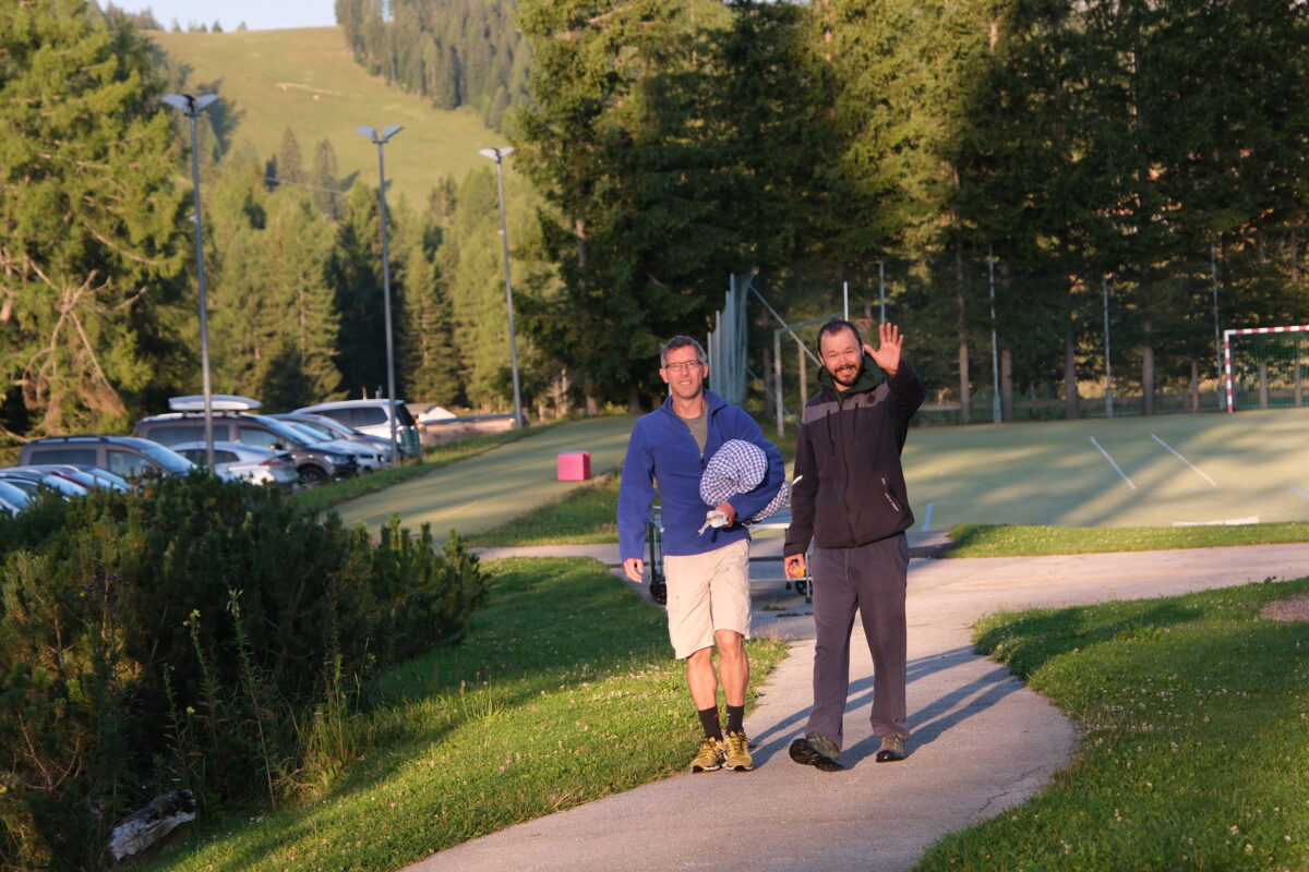 Die zwei Kursleiter gehen vom Parkplatz winkend einen Weg entlang.