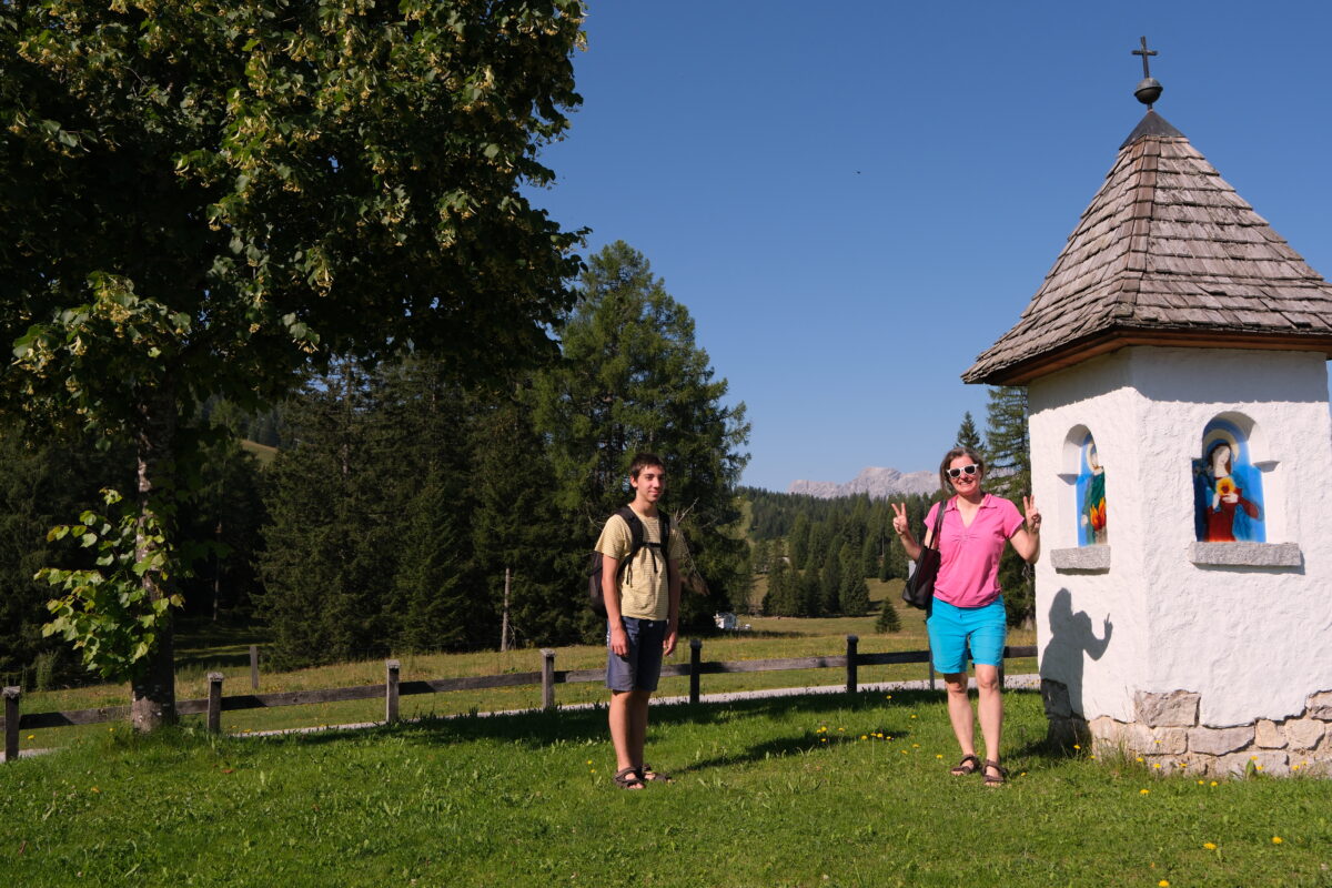 Mutter mit Sohn, die in der Natur spazieren gehen und bei einer kleinen Kapelle halt machen. Im Hintergrund die Bergwelt des Dachsteins.