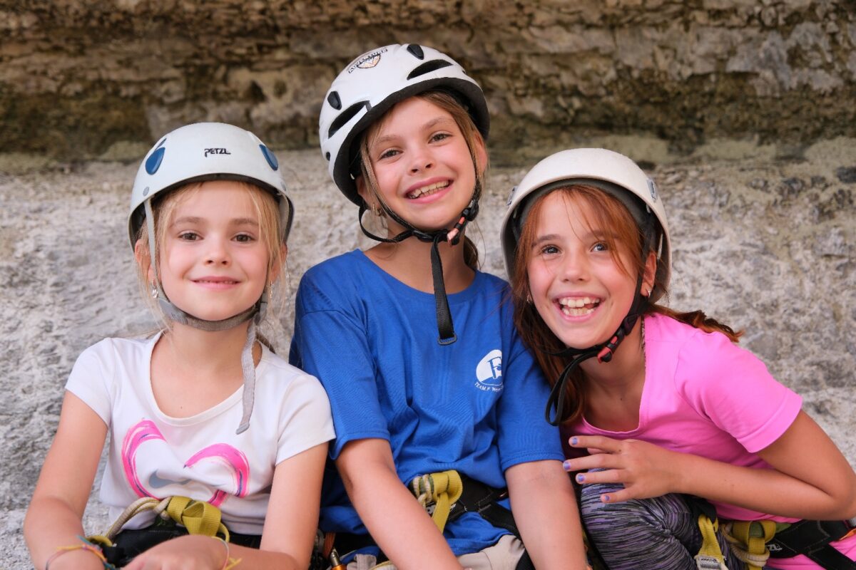 Fotopose von drei Mädchen im alter von etwa 12 Jahren. Sie tragen einen Kletterhelm und Klettergurte. Lachen fröhlich.