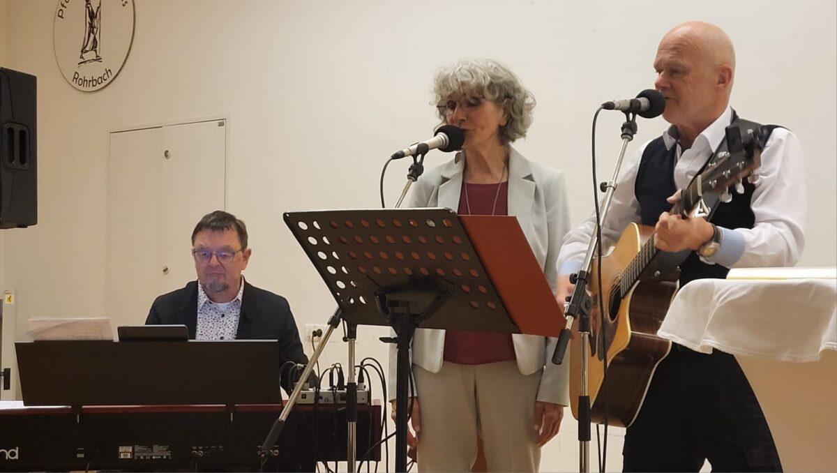 Ehepaar auf der Bühne, das ein Lied vorträgt. Die Frau singt und der Mann spielt zusätzlich Gitarre. Ein weiterer Mann im Hintergrund spielt Keybord.