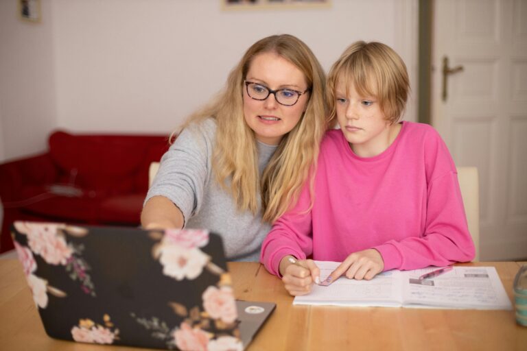 Mutter sitzt mit ihrer Tochter beim Tisch und lernt mittels Laptop mit ihr.