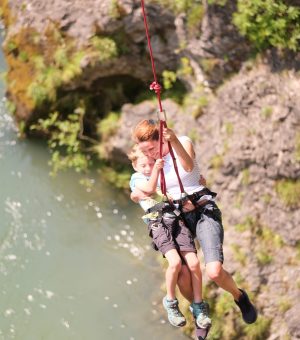 Imagefoto eines Bungeesprunges in eine Schlucht, unterhalb ist ein Gewässer. Frau ist mit einem Kind gemeinsam mit Gurten an einem Sprungseil gesichert.