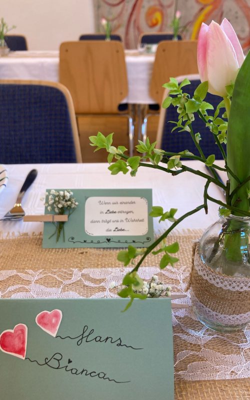 Tischdekoration aus einer Mitteldecke in Leinenoptik und weißer Spitze. Darauf eine Glasvase mit einer Tulpe und Grün. Namenskärtchen mit Herzchen darauf.