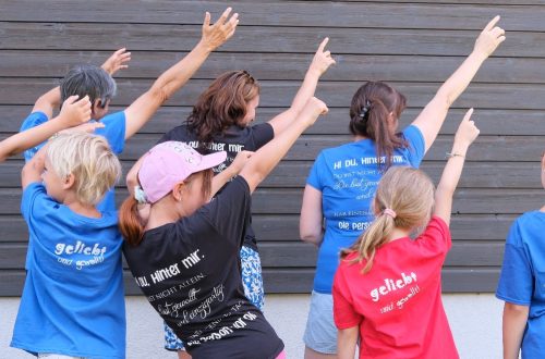 Gruppe von Kindern im Alter von 8 bis 14 Jahren und zwei erwachsenen Frauen, die mit dem Rücken zur Kamera stehen und T-Shirts präsentieren, die auf dem Rücken einen Ermutigungsspruch haben.